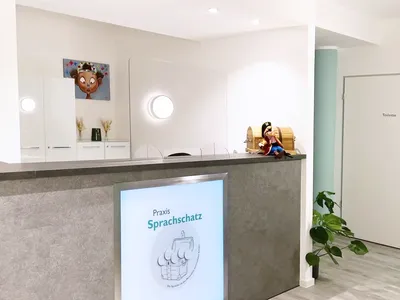Blick in den Eingangsbereich der Praxis auf die Theke mit Sprachschatz-Schild und dem Maskottchen, das auf der Ablagefläche sitzt. Ein türkiser Verlauf kommt von links in das Bild.