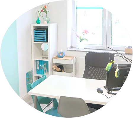 Leicht schräger Blick von oben auf einen Besprechungstisch mit Computer, zwei Stühlen und einem Hochstuhl für ein Kind. Die Sitzgruppe steht vor einem hellen Fenster mit zwei Regalen. Die Wand links ist türlkis.