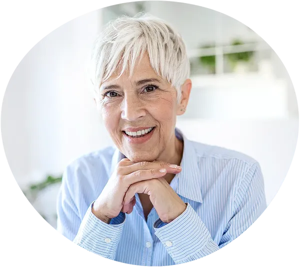 Sitzende grauhaarige Frau hat ein blaugestreiftes Hemand an und ihre Hände unter ihrem Kinn gefalten. Sie blickt zentral in die Kamera. Im Hintergrund sind ein weißes Regal und grüne Zimmerpflanzen zu sehen.