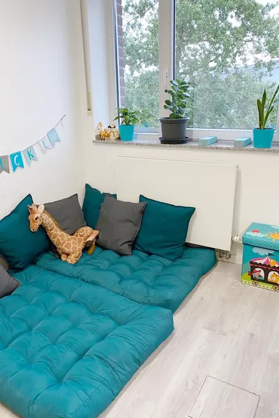 Spielecke mit grauen und türkisen Kissen auf dem Boden und Fähnchen an der Wand. Die Ecke befindet sich unter einem Fenster mit Heizung und einer Spielkiste im Vordergrund.