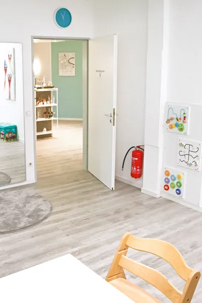 Blick aus einem Behandlungsraum in Richtung Flur. Im Vordergrund sieht man therapeutische Spielsachen an der Wand, im Flur blickt man auf eine türkise Wand.