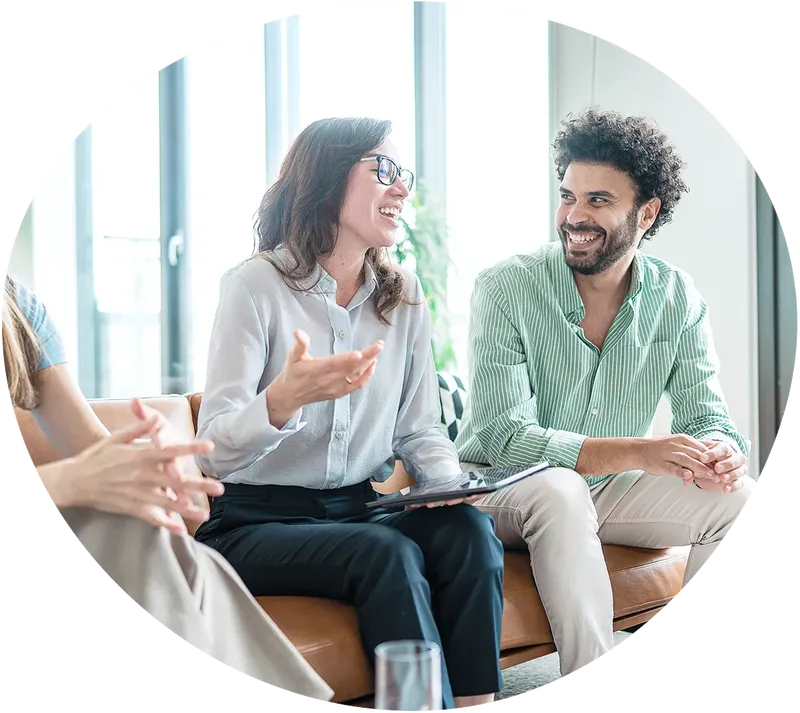 Drei Personen sitzen auf einer braunen Couch, die mittlere Frau erzählt und gestikuliert. Der Mann rechts neben ihr hört ihr aufmerksam zu und lacht sie an.